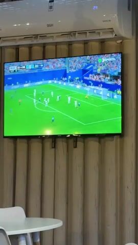 The video shows a television screen displaying a soccer match. The screen is mounted on a wall with vertical wooden paneling. Above the TV, an air conditioner is installed. In the foreground, the edge of a white table and a chair are visible. The soccer match shows players in white and blue uniforms on a green field with a goal on the right side of the screen. The score displayed on the screen indicates 00:00 and then changes to 00:03. The players appear to be in the middle of a play. At 00:04, the camera focuses on a player in a blue uniform with the number 20 on his back, who is holding his head. The air conditioner displays a temperature of 24.