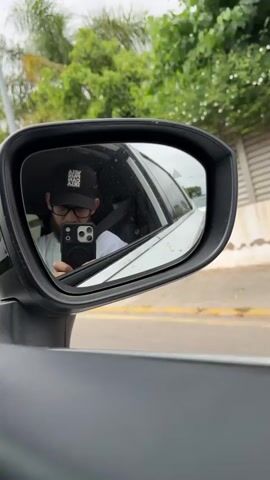 A person is seen in the reflection of a car's side mirror, wearing a black cap, black-framed glasses, and a white t-shirt. They have a beard and are holding a black smartphone, seemingly taking a picture or video of their reflection. A seatbelt is visible across their chest. The background visible outside the car in the mirror's reflection shows a road with a yellow line, green foliage, and distant structures under an overcast sky. The shot is taken from outside the car, focusing on the side mirror.