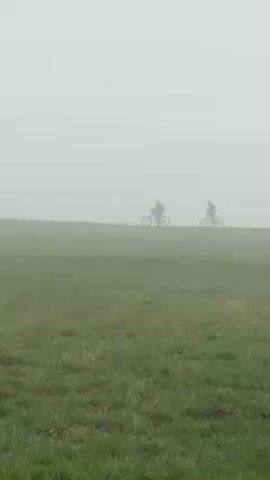 The video shows two people riding bikes across a grassy field on a foggy day. The figures are silhouetted against the dense fog in the distance. The foreground is a green field.