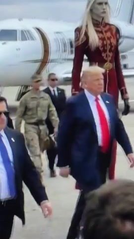 The media file depicts a scene on an airport tarmac with several individuals. In the foreground, three men in dark suits and sunglasses are walking towards the camera, appearing to be security personnel. One of them is wearing a blue tie, another a red tie, and the third a dark tie.

Behind them, to the left, is a white private jet with a gold stripe. Further back, a man in a military uniform walks away from the jet.

Dominating the center of the frame is a strikingly tall, slender figure with pale skin, long blonde hair, and sharp facial features. This individual is wearing an elaborate, long, dark red coat with gold and red embroidery, a gold belt, black gloves, and knee-high black boots. The figure appears to be a woman and has an imposing, almost regal presence.

In the mid-ground, behind the tall figure, other individuals in suits and military attire are visible, suggesting an official or important event. The overall atmosphere is formal and likely relates to an arrival or departure at the airport.

The lighting suggests it is daytime, possibly overcast, as there are no strong shadows. The setting is clearly an airport, indicated by the presence of the jet, tarmac, and other airport infrastructure in the background. No text is visible in the media file.