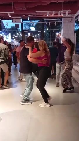 The video captures a group of people dancing in what appears to be an indoor venue. In the foreground, a man in a black t-shirt and light blue jeans is dancing with a woman in a pink top and black leggings. They are in a close embrace, suggesting partner dancing. The woman's right arm is raised, and her left hand is on the man's shoulder. The man's left arm is around her waist, and his right hand is on her back. Their bodies are angled, and their legs are in motion, indicating active dancing. In the background, other individuals are also dancing. To the right of the foreground couple, a woman with dark hair, wearing a patterned skirt and a light-colored top, is dancing. She has one arm raised. Further back, several other people are visible dancing in smaller groups. The environment is a well-lit indoor space with a high ceiling featuring exposed pipes and red accents. Large windows or glass doors are visible in the background, allowing some natural light to enter and showing an outdoor scene with trees and lights. The floor is a polished, light-colored surface. The overall atmosphere appears to be social and lively, consistent with a dance event or class. The video is a still image with no discernible motion or change occurring within the frame itself.