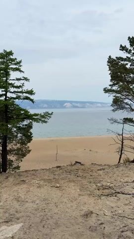 A scenic view of a beach on a lake with mountains in the distance. The beach is sandy and there are trees on either side of the frame. The lake is calm and the sky is overcast. There is a volleyball net set up on the beach. The mountains in the distance are snow-capped.