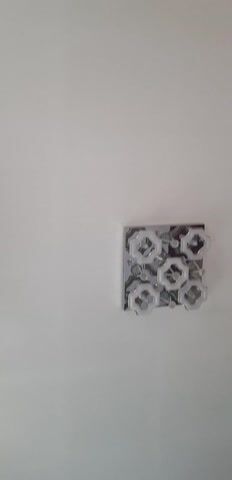 This media file contains a static shot of a modern, square ceiling light fixture mounted on a plain white wall. The light fixture has a metallic frame and features five hexagonal glass or plastic shades, each containing a bulb and a decorative element in its center. The overall design of the light is contemporary and geometric.

The lighting in the scene suggests it is daytime, as the wall is evenly illuminated with no harsh shadows that would indicate artificial light sources other than the fixture itself. The background is a simple, unadorned white wall, suggesting an indoor setting, possibly a room or hallway in a residential or commercial building. There are no people or other objects visible in the frame, focusing the viewer's attention solely on the light fixture. The environment appears clean and minimalist.

There is no discernible text within the media file. The overall mood is neutral and functional, highlighting the design of the light fixture against a blank backdrop. The setting does not provide specific clues about the time of day beyond it being lit, nor does it offer any indication of weather conditions. The location context of Kyzyl-Saray, Kyrgyzstan, is external information and not visually represented within the image itself.