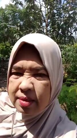 A close-up video shows a middle-aged woman wearing a light-colored hijab and a light brown shirt. Her right arm is raised, with her hand resting on the top of her head. She is looking directly at the camera with a slight smile. The background is blurred but appears to be an outdoor setting with trees and a wooden structure. The lighting suggests it is daytime.