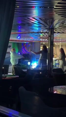 Three women are dancing and having fun in a dimly lit room with colorful lights. One woman in the foreground is wearing a white hoodie and pants and is dancing with her arms up. Another woman in the middle is wearing a blue top and gray pants and is dancing energetically. A third woman in the background is wearing a white dress and is also dancing. The room appears to be a lounge or club with seating and a bar in the background. The lights are flashing and creating a party atmosphere.