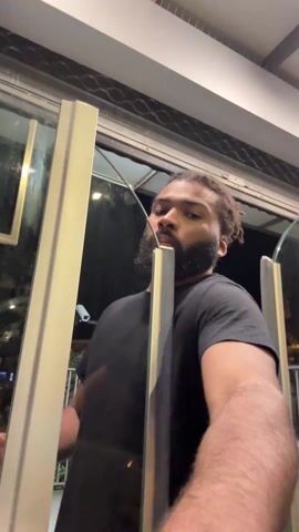 A man is seen from a low angle, climbing over turnstile-like barriers at night. He wears a black t-shirt, black pants, and slides. The barriers are silver-colored with glass panels. Outside, there is a street with a security camera and a ticket vending machine visible. The man places his hands on the barriers to pull himself over.