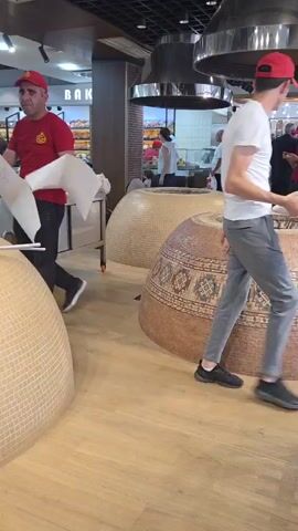 A man wearing a white t-shirt and grey pants, with a red baseball cap on his head, enters the frame from the right holding a round, tan-colored object. He walks towards the center of the room where two large, dome-shaped clay ovens are visible. The oven in the foreground is decorated with a patterned mosaic of geometric shapes in brown, beige, and colorful squares. The oven behind it appears to be lighter in color and has a textured surface. Several other people are in the background, some appearing to be customers or employees. One person in a white chef's hat is seen working behind a counter. The room has a light-colored wooden floor and industrial-style pendant lights hanging from the ceiling. The man holding the object walks past the foreground oven and disappears behind it. The scene appears to be a bakery or food establishment.