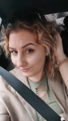 A woman with curly reddish-blonde hair is sitting in a car, wearing a tan pinstripe blazer, a green shirt with cutouts, and a bracelet on her left wrist. A seatbelt is fastened across her chest. She is looking directly at the camera and smiling slightly. The background appears to be an urban environment with other vehicles visible through the car window.