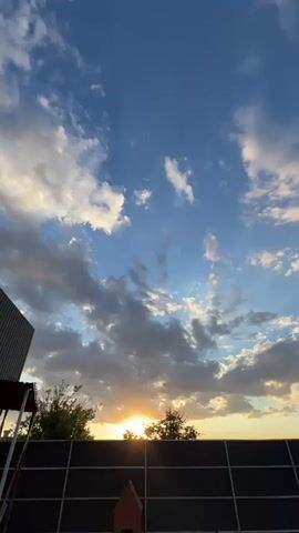 This is a vertical, low-angle shot of a sky at sunset. The sun is low on the horizon, peeking through the trees and casting a golden glow. Clouds are scattered across the blue sky, some white and fluffy, others darker and more dramatic. In the foreground, a dark fence runs across the bottom of the frame. To the left, a sliver of a building is visible. The overall impression is one of natural beauty and tranquility, with the setting sun creating a warm and inviting atmosphere.