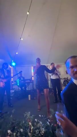 This is a video of a band performing on a stage at an indoor event, likely a wedding reception or party. A female singer in a short, sparkly black dress is at the forefront, singing into a microphone. To her left, a male guitarist in a suit plays his instrument. Behind the singer and guitarist, a keyboardist is seated at an electronic keyboard. To the singer's right, a male vocalist in a white shirt and black pants sings into a microphone, and further to the right, a drummer is playing a drum set. The stage is lit with various colored lights, including red, green, blue, and purple. The band is performing under a large, white tent or draped ceiling, with string lights illuminating the space. In front of the stage, the audience is visible, with some people dancing and others observing. The audience is dressed in party attire, with women in dresses and men in shirts and suits. Some people are holding drinks. The overall atmosphere appears festive and energetic.