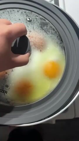 The video shows a close-up, overhead view of a person's hand lifting a glass lid from a frying pan. Inside the pan, two fried eggs with intact, bright orange yolks are visible, along with three browned sausages. The eggs are surrounded by a thin layer of cooked egg whites. The pan is dark and appears to be on a stovetop, with hints of a white surface in the background. The lid has a gray silicone rim and a black knob. The video is shot in a dimly lit environment.