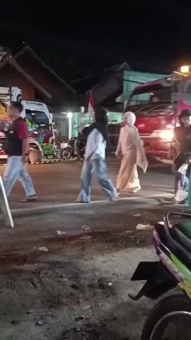 A group of people, some wearing hijabs, are standing on a road at night. A large red truck with a blue tarp covering its bed is stopped on the road. The truck appears to be an industrial vehicle, possibly a dump truck or a flatbed. In the background, another truck with colorful graphics is visible, as well as some buildings and streetlights. A motorcycle is parked in the foreground. The ground is dirty and littered with debris. The people appear to be interacting, possibly talking or observing something. The overall scene suggests a casual gathering or an event taking place on the roadside at night.