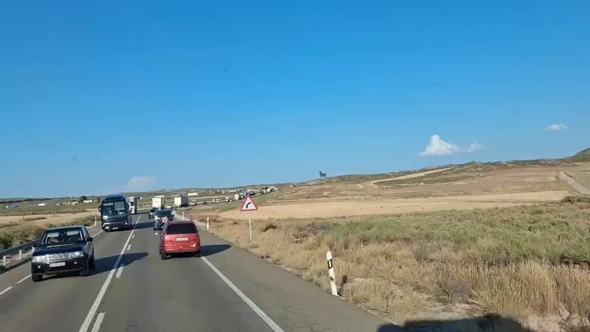 The video shows a view from a moving vehicle on a highway. The road is asphalt with white lane markings. There are several vehicles on the road, including a black bus, a red car, and numerous trucks. The trucks appear to be in a long line, suggesting traffic congestion. To the right of the road, there is dry, grassy terrain with some bushes and a few scattered trees. In the distance, on top of a hill, stands a large silhouette of a bull. The sky is clear and blue. A traffic sign indicating a sharp curve to the right is visible on the side of the road. The overall impression is a rural highway scene with a significant traffic jam and a prominent landmark in the distance.