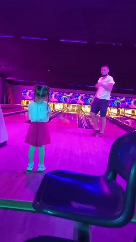 This is a video clip showing a father and daughter at a bowling alley. The scene is illuminated by purple and pink neon lights, creating a vibrant and energetic atmosphere typical of a bowling alley. The father, wearing a white t-shirt and dark shorts, is standing on the bowling lane, and the daughter, in a white top and red skirt, is on the approach. The daughter appears to be excited and is gesturing with her hands. The bowling alley features multiple lanes with pins at the end and a ball return system. In the foreground, a dark blue chair is visible, suggesting the viewer is seated. The video also briefly shows a rack of colorful bowling balls, including green, yellow, orange, and blue ones. The overall impression is a fun family outing, likely taking place in the evening given the lighting. No visible text is present in the clip.