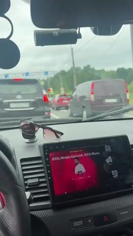 The video begins inside a car, looking forward through the windshield. The driver's side of the steering wheel and dashboard are visible on the left, with a portion of the infotainment screen in the center displaying album art for "JEEG, Igor' Krutoy, JEEG Music." A pair of sunglasses rests on a stand on the dashboard. Outside, traffic is heavy, with several cars and a van visible on a cloudy day. The camera appears to be mounted on the dashboard or windshield. The scene suggests the car is stationary or moving slowly in traffic.
