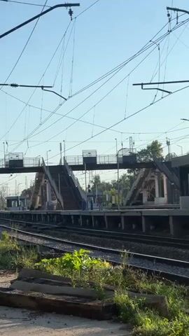 The video shows an outdoor view of a train station platform with railway tracks in the foreground. Overhead power lines crisscross the sky, which is clear and blue. On the platform, there are two staircases leading up to a pedestrian overpass that spans the tracks. A small building, possibly a ticket booth or control room, is visible on the right side of the platform. Two individuals are walking on the platform in the distance, and some greenery grows along the edge of the tracks in the foreground. The scene appears to be set during the daytime, likely in the morning or late afternoon, given the angle of the sunlight and shadows.
