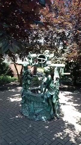 The video displays a bronze sculpture of a well in a garden setting. The sculpture features a woman sitting on the edge of the well, with her head tilted back and her eyes closed. She is adorned with flowing drapery and appears to be in a contemplative or serene pose. The well itself is intricately designed with detailed carvings and motifs. Above the well, a wooden crank mechanism is visible, suggesting the functionality of the well. Two cherubic figures are also part of the sculpture, positioned near the crank, adding a whimsical or angelic element. The environment surrounding the sculpture is lush with greenery, including trees with dark red leaves and foliage, and the ground is paved with brickwork, creating a patterned walkway. Sunlight filters through the trees, casting dappled shadows on the scene, contributing to a peaceful and somewhat mysterious atmosphere. The video pans slowly across the sculpture, offering a 360-degree view.