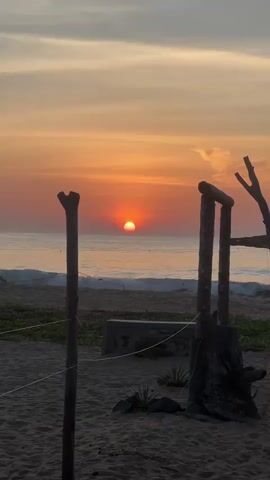 This video shows a sunset over the ocean. The sun is a bright orange orb, partially submerged in the water. The sky is a gradient of orange, pink, and yellow, with some wispy clouds. In the foreground, there is a sandy beach with some driftwood structures. A sign that says "YOGA" is visible on one of the structures. The waves are gentle, lapping against the shore. The scene is serene and peaceful.