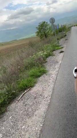 The video shows a view from a moving vehicle, likely a car, on a rural road. The road is paved, with a gravel shoulder on the side visible to the camera. Beyond the shoulder, there is grassy vegetation and some small trees. In the distance, rolling hills or mountains are visible under a cloudy sky. The color of the vegetation suggests it is likely springtime. A cultivated field can be seen beyond the roadside vegetation. Road signs are visible but not clearly legible due to the movement and distance. The overall impression is of a scenic drive through a rural area.