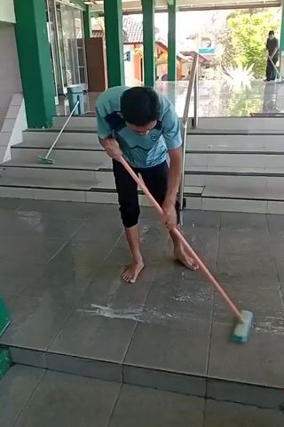 A young person, barefoot and wearing a teal and black t-shirt and black pants, is mopping a tiled floor with a mop. The floor appears wet and soapy. The person is standing on the lower level of a structure with green pillars and a white roof. In the background, there are stairs leading up, and on the top level, another person is visible standing near a railing. The environment appears to be an outdoor covered area, possibly a mosque or public building, with sunlight filtering through. There is a broom leaning against the stairs on the left side of the frame.