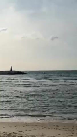The video shows a beach scene with a calm ocean under a cloudy sky. There is a breakwater extending into the water on the left side of the frame, and a lighthouse stands at its end. Several buoys are visible in the distance. The waves are gentle, and the sand is visible in the foreground.
