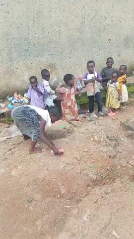 In Kagarko South, Nigeria, a group of children gathers near a textured, pale wall. A woman in a black and white striped outfit bends over, appearing to interact with something on the ground. To her right, a group of young children stand and watch. Among them, a boy in a purple and white shirt and dark pants is holding a stick. Another child in a patterned dress is to his left, and further to the right, children in yellow and white dresses are visible, with one child in a dark shirt standing behind them. The ground is a mix of dirt and rocks, with some debris scattered around. The lighting suggests it is daytime, with no clear indication of the time of day. The scene appears informal and likely outdoors. There is no visible text in the media file.