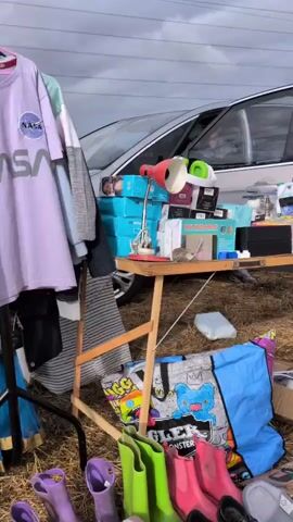A silver car is parked on a grassy field with its doors open. A table is set up in front of the car, displaying various items for sale, including boxes, a lamp, and bags. The ground around the table is covered with more items, such as clothes, boxes, and bags. Another car, a black Nissan, is parked to the right of the silver car. A person is visible sitting in the driver's seat of the silver car. The sky is overcast.