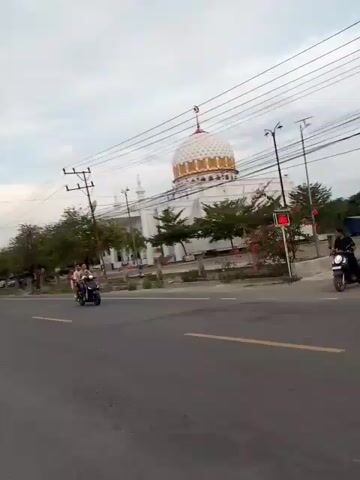 The video shows a mosque with a white dome and gold accents. The mosque is surrounded by trees and utility poles. Several vehicles, including a white car, a scooter, and a black truck, are driving by on a road in front of the mosque. The sky is overcast.