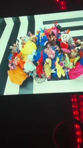 The video shows a recording of a screen displaying a montage of flamboyant and colorful fashion scenes. In the first clip, two figures with elaborate hair and makeup pose against a red background. The subsequent shots showcase different models in eccentric outfits, including corsets, large hats with feathers, and avant-garde designs, with many of them dancing and posing in front of striped and solid-colored backgrounds. The final scene features two figures in full-skirted gowns against a red backdrop, mirroring each other's poses. The room in which the recording is taking place is dark, with red string lights visible on the right side.