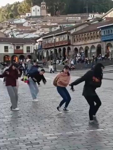 The video shows a group of people dancing in a plaza or town square. Most of the dancers are young women. One dancer is twirling a long dark object, possibly a whip. Other dancers hold what appears to be a colorful flag. Spectators are watching from the sides, some seated. The plaza is paved with cobblestones. Buildings with tiled roofs surround the plaza. In the background, a hill with houses is visible. The sky is overcast.