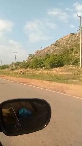 The video captures a view from a vehicle, likely from the perspective of a passenger looking out the side window. A cracked side-view mirror is prominently in the foreground, reflecting a blue object and part of the interior of the vehicle. Beyond the mirror, a paved road stretches into the distance, bordered by a dry, grassy shoulder. In the background, a rocky, treeless hillside rises against a pale blue sky with scattered white clouds. A utility pole with wires is visible on the left side of the road. The overall scene suggests a rural or semi-arid environment during the daytime.