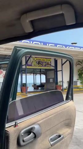 The video captures a view from inside a vehicle, likely a car, in Puerto La Cruz, Venezuela. The primary focus is an outdoor, open-air structure or pavilion.

A large, yellow banner is prominently displayed at the entrance of the structure. It reads "¡DESCARGA Y GANA YA!" (Download and Win Now!) and advertises "APUESTAS ROYAL." The banner features various categories with accompanying images, including "ANIMALITOS" (Animals), "HIPISMO" (Horse Racing), "MINIJUEGOS" (Mini-games), and "APUESTAS DEPORTIVAS" (Sports Betting). Two QR codes are visible, along with the website address "WWW.APUESTASROYAL.COM" displayed twice.

Beyond the banner and within the open structure, there are tables and chairs, indicating a seating or dining area. Further back, a metal railing overlooks a large body of water, presumably the Caribbean Sea, under a hazy daytime sky. Potted plants are positioned on either side of the open space.

Briefly, between 0:02 and 0:04, the camera pans inside the vehicle, showing a person's leg in light-colored pants and a car floor mat with "CORSA" embossed on it. The video then returns to the exterior view of the building and banner. Upbeat music with Spanish lyrics can be heard playing in the background throughout the clip.