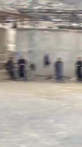 The video shows a group of people in an outdoor area. Some are sitting on a concrete structure, while others are standing around. The area appears to be somewhat under construction or in a state of disrepair, with exposed brick and concrete. There is a metal structure overhead. The background shows a building on a hillside. The atmosphere is casual, with people seemingly observing something out of frame.