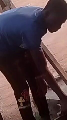 A person is sweeping. They are wearing a blue shirt, dark pants with some embroidered detailing, and a wristband. The person is using a straw broom to sweep debris from a sunken area under a wooden structure. The environment appears to be outdoors with a dirt or sandy surface. A motorcycle wheel can be seen briefly in the background.