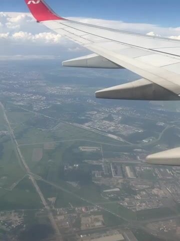 The image is an aerial view from an airplane window, looking out over a landscape. The primary subject is the wing of the airplane, which occupies the upper right portion of the frame. It is white with a red logo at the tip, featuring a stylized "N" or "V". Below the wing, a vast expanse of land unfolds.

The scene depicts a transition from rural to urban or industrial areas. In the foreground, there are agricultural fields with distinct patterns of cultivation. Further into the distance, buildings and infrastructure become more prominent, suggesting a developing urban or industrial zone. There are numerous structures, roads, and what appear to be factories or industrial complexes with multiple buildings. Green spaces and patches of trees are interspersed throughout the landscape.

The sky is blue with scattered white clouds, indicating fair weather. The lighting suggests it is daytime, possibly late morning or afternoon, given the brightness. The perspective from the airplane provides a sweeping view of the terrain, revealing the layout of settlements and infrastructure from above. No people are visible in the image. The overall mood is one of travel and observation, a detached view of the world below.

No legible text is present in the image, other than the logo on the airplane wing. The location context provided indicates the scene is over Stolbishche, Russia.