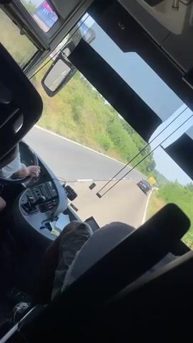 The video provides a first-person perspective from inside a bus. The camera is angled to show the driver's area, the dashboard, and the front windshield, through which a winding road is visible. The road is bordered by green, vegetated hills. Two vehicles are seen on the road ahead. The camera then pans to the interior of the bus, revealing rows of seats occupied by passengers. Most passengers appear to be children or young adults, some of whom are looking around or resting. The overall impression is of a group traveling by bus on a sunny day, likely on a road trip or excursion.