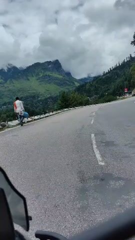 The video shows a first-person perspective of someone riding a motorcycle on a paved road through a mountainous, forested area. The road curves to the right, with a steep, rocky, and tree-covered mountainside to the right and a lower, also tree-covered slope to the left. The sky is overcast with dramatic clouds. There are white lines on the road, marking lanes or shoulders, and a white guardrail along the left side of the road. A red sign is visible on the right side of the road, and further up the mountain, a few buildings or structures can be seen. The front of the motorcycle, including the windshield and handlebars, is visible in the foreground.