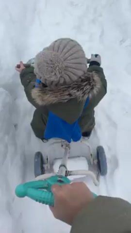 A person is using a child's stroller tricycle to push through a snowy path next to a red car. The child, wearing a winter hat, coat, and scarf, is sitting on the trike and tries to grab the snow with their hands. The scene appears to be in a residential area after a snowfall.