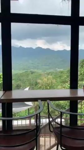 Here's a description of the scene in the video:

The video presents a scenic view from inside a room with a large window. The primary focus is the landscape visible through the window, which includes lush green hills and a mountain range in the distance under a cloudy sky. 

In the foreground, there's a wooden countertop running along the window, paired with a few dark-colored, modern bar stools.  Outside, roofs of nearby buildings are seen. The room itself has a black metal frame window structure, and a white rope hangs from the ceiling. The overall environment appears to be a cafe or restaurant with an emphasis on the view.