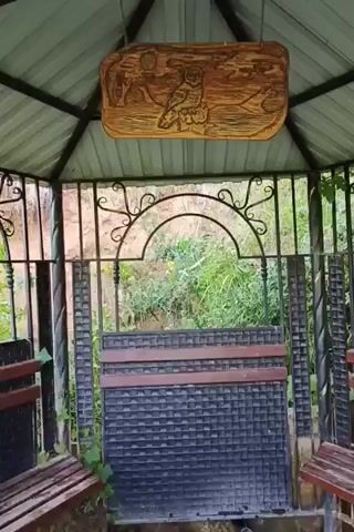 A video pans across the interior of a gazebo. The gazebo has a metal roof with a hexagonal frame, and wooden benches are attached to the sides. In the center of the gazebo, a decorative metal grate separates the space, and behind it, a small fountain flows. The fountain consists of a pipe from which water streams into a rocky basin. The area around the fountain is muddy, and a small stream of water flows away from the basin. The background shows a hillside covered in greenery. A carved wooden sign hangs above the grate, depicting an owl.