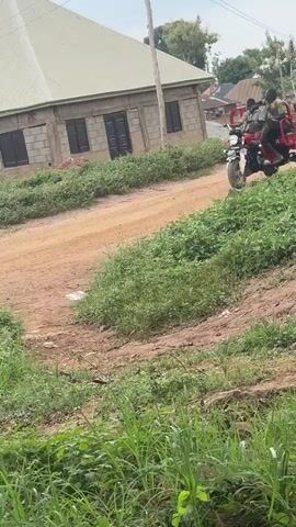 A red three-wheeled motorcycle with a cargo bed is driving on a dirt road. Two people are riding on the motorcycle, one driving and one passenger. The motorcycle is in the middle of the frame. In the background, there is a building under construction and some trees. The sky is overcast.