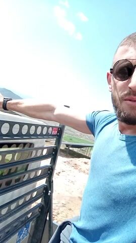 A man with a beard and sunglasses is standing on a hill with a solar panel farm in the background. The sky is blue with some clouds. He is wearing a blue t-shirt and a silver necklace. He is holding onto a black metal structure. He turns his head from side to side and smiles.