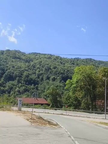 The video shows a roadside scene with a blue sky and green forested hills in the background. A paved road curves through the foreground. On the left, there is a white sign and a red-roofed building. Several streetlights are visible along the road. In the middle ground, there are trees and more buildings with red roofs. A silver car is seen driving on the road in the distance on the left. A large green tree stands prominently in the foreground on the right. The video is a static shot, with no apparent camera movement.