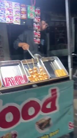 The video begins with a close-up of a street food vendor preparing skewers of hot dogs over a grill. The vendor, a young man wearing a dark hoodie, is visible behind a cart laden with food items. The cart has a teal-colored banner with red text that is partially obscured, but appears to say "food". Several trays of food are arranged on the cart, including skewers of sliced hot dogs and whole hot dogs. In the background, more vendors and shoppers are visible, along with stalls selling various goods, including toys. The scene appears to be a night market. The camera pans to show more of the market, including a variety of colorful toys displayed on stalls. People are seen walking through the market and browsing the goods. The video ends with a shot of a sign that says "Kebab".
