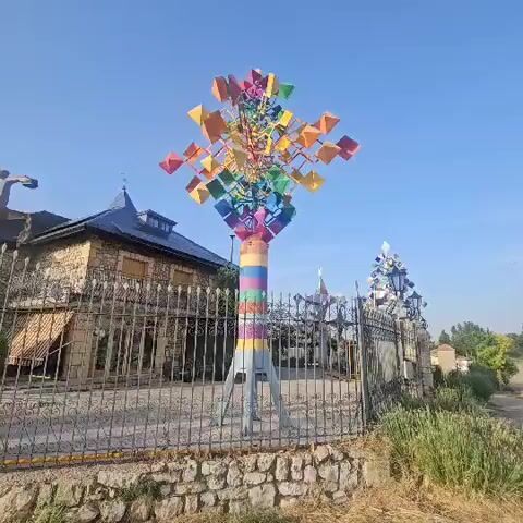 The video shows a close-up of a colorful, kinetic sculpture that resembles a large pinwheel or windmill. The sculpture is made of many small, square, and diamond-shaped pieces in various colors of the rainbow, attached to a central pole. The pole itself is also painted with colorful horizontal stripes. The sculpture is outdoors, with a clear blue sky in the background. To the left of the sculpture is a building with a dark-tiled roof and stone facade, behind a black metal fence. Other similar, smaller sculptures can be seen in the background behind the fence. The ground in front of the fence is covered with loose stones and some dried vegetation. The overall impression is one of a whimsical and artistic outdoor display.