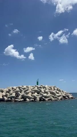 The video shows a person taking photos from a boat on a sunny day. The camera starts focused on a large pile of concrete shapes in the water, with a green navigational light on top. The view pans right along a breakwater, then shows the perspective from the boat, with a man in a hat and striped shirt taking photos with his phone towards the shore. Other people are also visible on the boat. The sky is blue with puffy white clouds, and the water is a bright, clear turquoise. The boat appears to be moving away from the land.