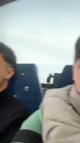 The video appears to be taken on a moving bus. The first shot features a young man with short dark hair wearing a black zip-up sweater. The camera pans to a young woman in a black leather jacket who covers her face with her hand. The camera then shows a young woman in a pink puffer jacket who leans back in her seat. The final shot shows a young man with curly brown hair in a beige puffer jacket. The bus has blue patterned seats and a large window showing a blurry outdoor view.