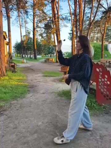 A young woman with long dark hair and glasses stands in a dirt path outdoors. She wears a dark striped shirt and light gray pants with white sneakers. She is holding a small object in her right hand, which she extends upward. The background shows tall trees and some buildings.