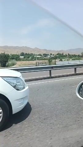 A white car is traveling on a highway. A white minivan passes the car on the left. The car continues to travel on the highway past trees and mountains in the background. The sky is blue with some clouds.
