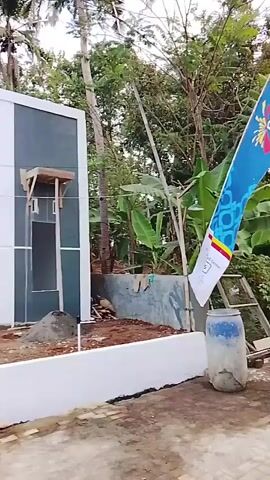 The video shows a newly constructed or renovated house with a modern design. The exterior walls are white with dark gray accents. There are several doors and windows. In front of the house, there is a pile of sand, some gardening tools, and a partially constructed white wall or planter box. A person wearing a blue shirt and an orange safety vest is sitting on the white wall. Another person in a light-colored shirt and dark pants is standing and talking on a phone. A red and black motorcycle is parked on the paved area in front of the house. A silver car is parked on the driveway. The surroundings include trees and foliage, indicating an outdoor environment. The sky is visible and appears to be cloudy or overcast. The overall scene suggests a construction or property viewing context.